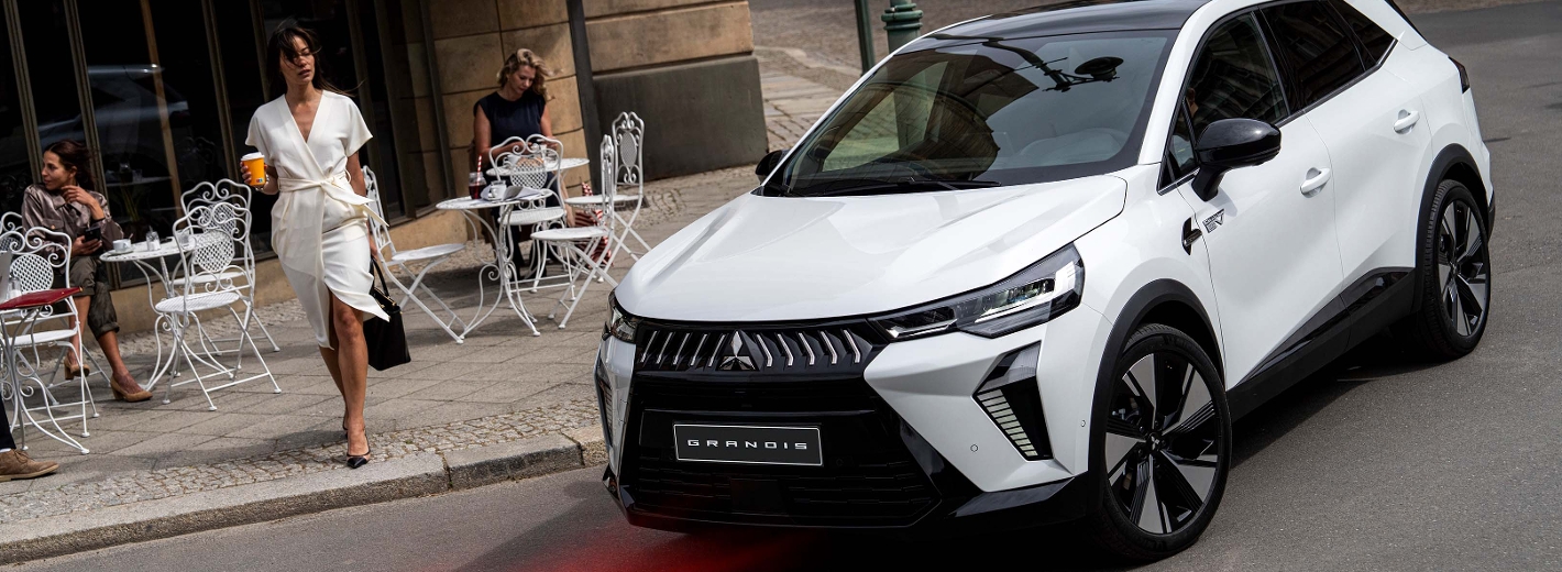 weißer Mitsubishi Grandis fährt an einem Straßen-Café mit weißen Tischen und Sesseln vorbei und eine Frau im weißen Kleid steht am Straßenrand.