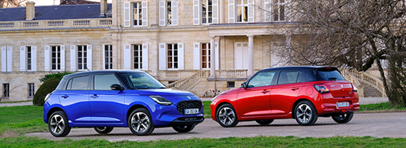 Zwei Fahrzeuge des Modelles Suzuki Swift stehen einander gegenüber. Das linke Auto in Blau, das rechte Auto in der Farbe Rot. Sie parken vor einem renovierten alten Gebäude, ein Schloss.