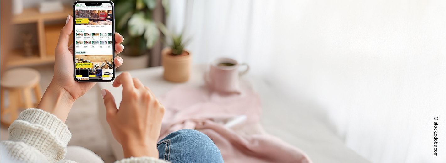Eine Person sitzt gemütlich auf einem Sofa und hält ein Smartphone in der Hand. Auf dem Display ist der Onlineshop von lagerhaus.at zu sehen. Die Person trägt eine helle Strickjacke und Jeans. Im Hintergrund stehen eine kleine Pflanze, eine Tasse und weiche Wohntextilien, wodurch die Szene warm und entspannt wirkt.