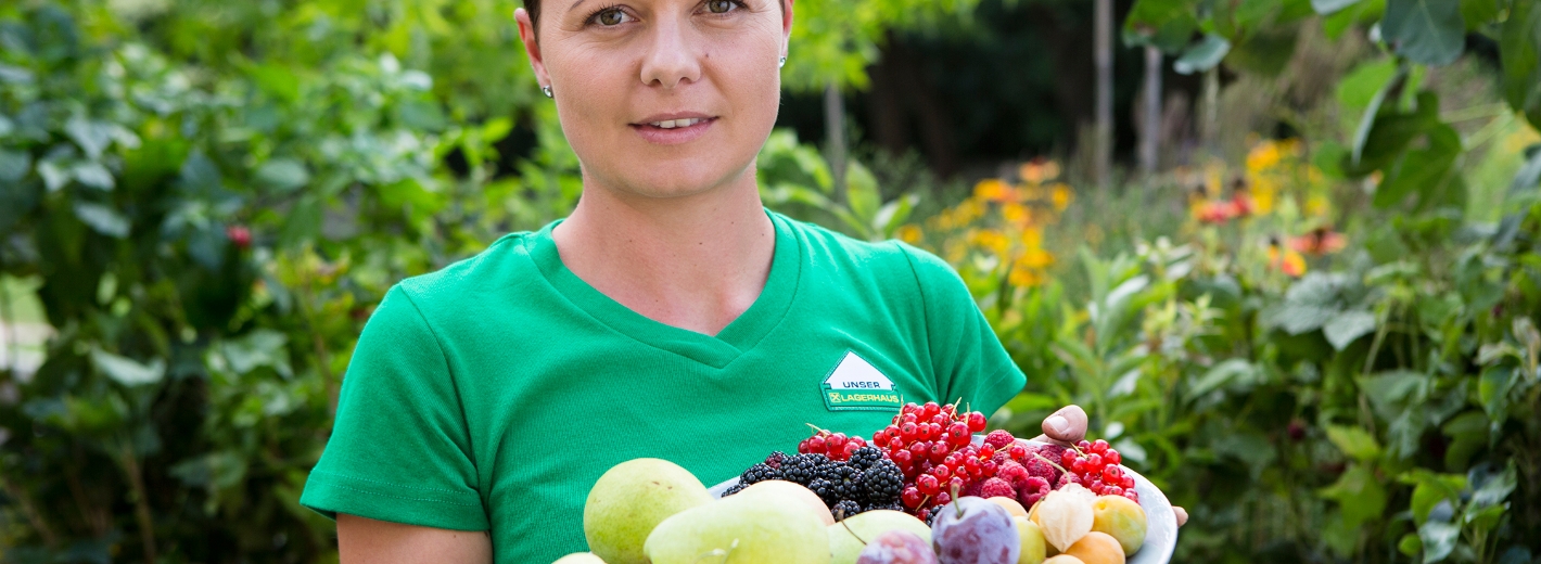 Frau mit grünem Lagerhaus-T-Shirt hält ein Teller mit frischem Obst in der Hand.