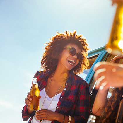 Lachende junge Frau mit Sonnenbrille und Flasche in der Hand vor einem Kleinbus