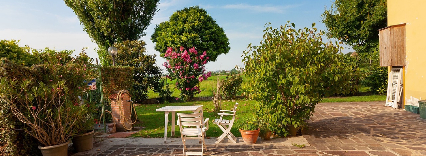 Schöner Garten mit Sträuchern und Bäumen, Sitzgelegenheit und terrakottafarbenem gepflasterten Boden vor einer gelben Hausmauer