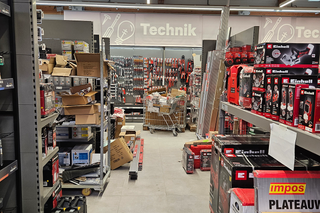 In einem Bau- und Gartenmarkt stehen lange, hohe Regale in mehreren Reihen. Die Regale sind aus Metall und reichen fast bis zur Decke. Darin befinden sich viele unterschiedliche Produkte in Kartons, Kunststoffverpackungen oder auf Haken aufgehängt. In einem Bereich stehen Farbeimer, Lacke und Pinsel. In einem anderen hängen Werkzeuge wie Hämmer, Schraubendreher und Sägen. Weiter hinten sind Gartengeräte wie Blumentöpfe, Erde, Dünger und Gießkannen gestapelt. Die Gänge zwischen den Regalen sind breit und gut begehbar. Über den Regalen hängen Schilder, die die verschiedenen Warengruppen anzeigen.