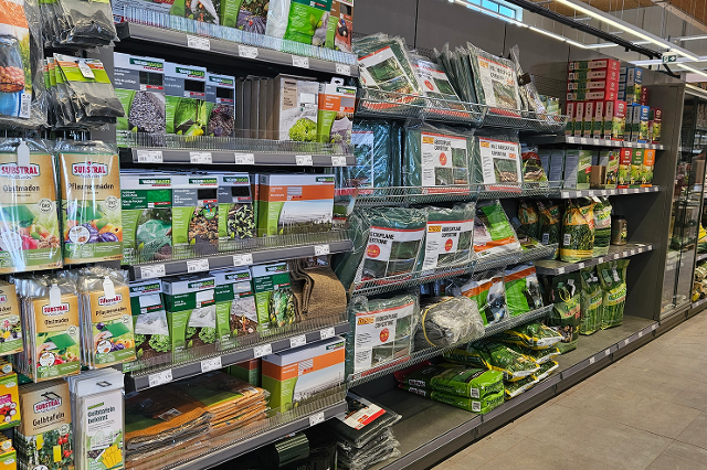In einem Bau- und Gartenmarkt stehen lange, hohe Regale in mehreren Reihen. Die Regale sind aus Metall und reichen fast bis zur Decke. Darin befinden sich viele unterschiedliche Produkte in Kartons, Kunststoffverpackungen oder auf Haken aufgehängt. In einem Bereich stehen Farbeimer, Lacke und Pinsel. In einem anderen hängen Werkzeuge wie Hämmer, Schraubendreher und Sägen. Weiter hinten sind Gartengeräte wie Blumentöpfe, Erde, Dünger und Gießkannen gestapelt. Die Gänge zwischen den Regalen sind breit und gut begehbar. Über den Regalen hängen Schilder, die die verschiedenen Warengruppen anzeigen.
