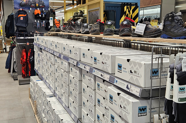 In einem Bau- und Gartenmarkt stehen lange, hohe Regale in mehreren Reihen. Die Regale sind aus Metall und reichen fast bis zur Decke. Darin befinden sich viele unterschiedliche Produkte in Kartons, Kunststoffverpackungen oder auf Haken aufgehängt. In einem Bereich stehen Farbeimer, Lacke und Pinsel. In einem anderen hängen Werkzeuge wie Hämmer, Schraubendreher und Sägen. Weiter hinten sind Gartengeräte wie Blumentöpfe, Erde, Dünger und Gießkannen gestapelt. Die Gänge zwischen den Regalen sind breit und gut begehbar. Über den Regalen hängen Schilder, die die verschiedenen Warengruppen anzeigen.