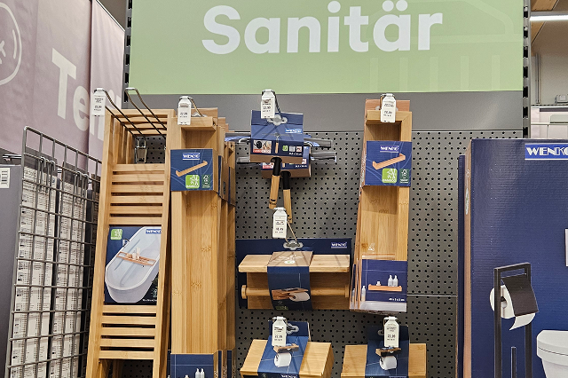 In einem Bau- und Gartenmarkt stehen lange, hohe Regale in mehreren Reihen. Die Regale sind aus Metall und reichen fast bis zur Decke. Darin befinden sich viele unterschiedliche Produkte in Kartons, Kunststoffverpackungen oder auf Haken aufgehängt. In einem Bereich stehen Farbeimer, Lacke und Pinsel. In einem anderen hängen Werkzeuge wie Hämmer, Schraubendreher und Sägen. Weiter hinten sind Gartengeräte wie Blumentöpfe, Erde, Dünger und Gießkannen gestapelt. Die Gänge zwischen den Regalen sind breit und gut begehbar. Über den Regalen hängen Schilder, die die verschiedenen Warengruppen anzeigen.