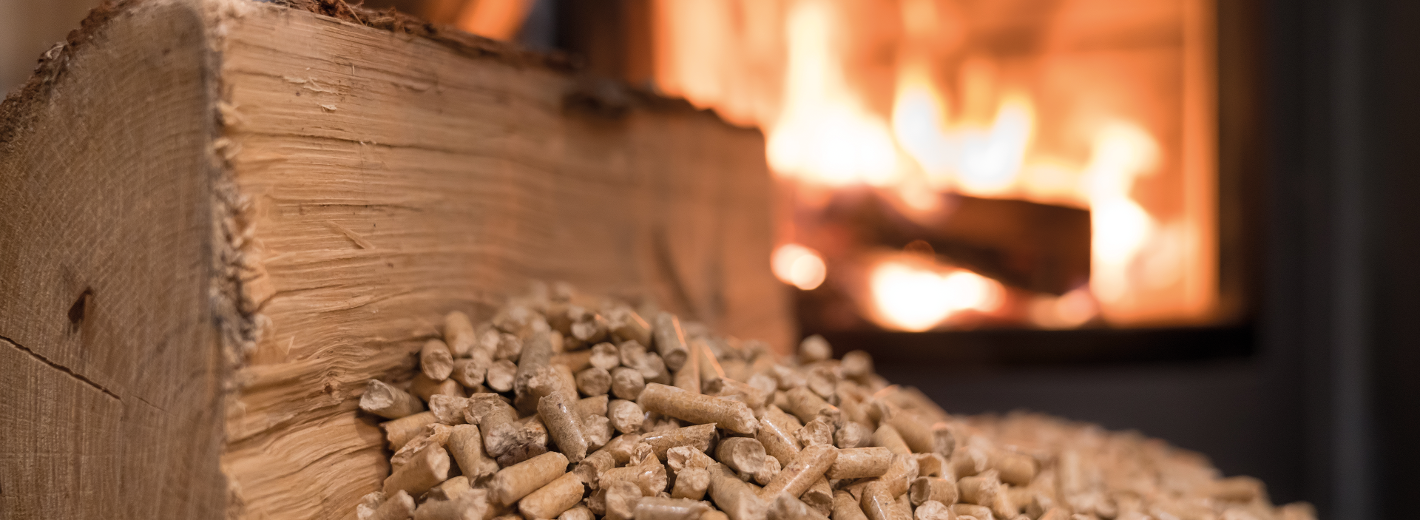 Ein gemütlicher Kamin mit loderndem Feuer im Hintergrund. Davor liegt ein großer unbehandelter Holzblock, vor dem ein Haufen heller Holzpellets auf dem Boden ausgeschüttet ist – Symbol für die Wärmequelle aus natürlichem Rohstoff.
