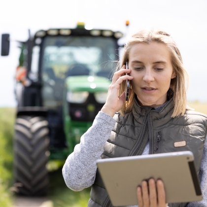 Eine Frau mit blonden Haaren telefoniert konzentriert mit einem Smartphone, während sie gleichzeitig ein Tablet in der anderen Hand hält. Sie trägt eine graue Steppweste über einem hellen Oberteil. Hinter ihr ist unscharf ein grüner Traktor auf einem Feld vor hellem Himmel zu erkennen.