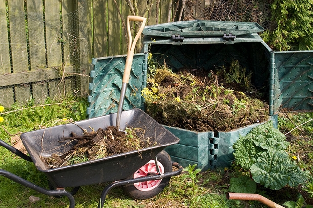 Geöffneter Kunststoffkomposter im Garten mit Gartenabfällen, davor Schubkarre und Spaten auf einer Wiese.