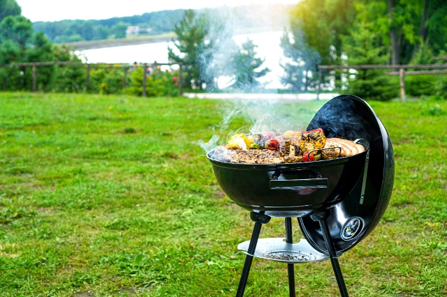 Kugelgrill mit offenem Deckel auf einer grünen Wiese, bestückt mit Fleisch und Gemüse über glühender Holzkohle