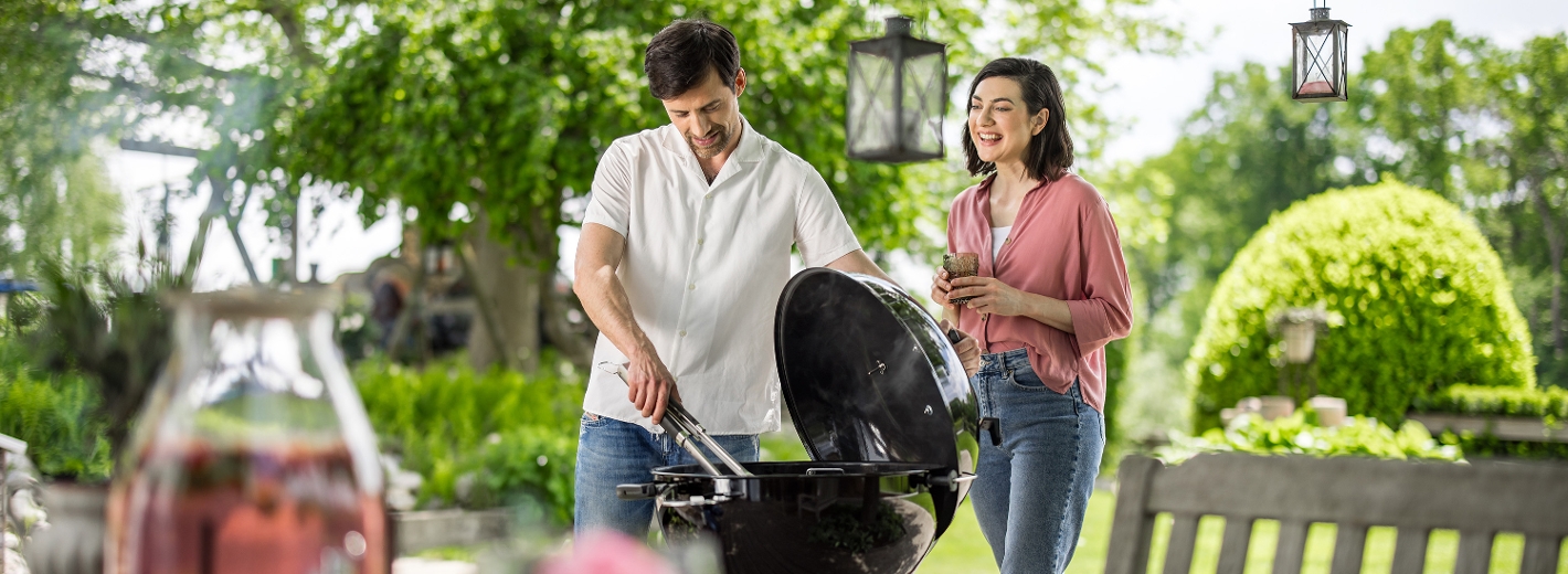 Sommerliche Grillszene im Garten: Ein Mann grillt am Kugelgrill, eine Frau steht neben ihm