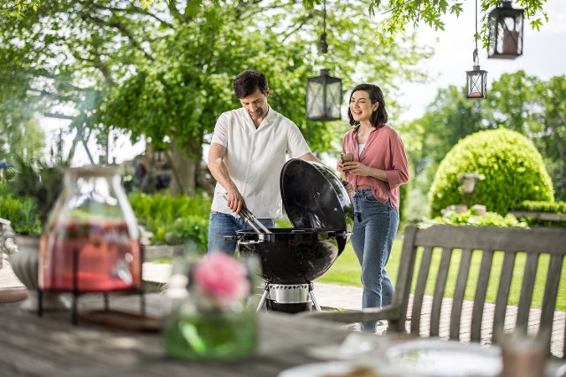 Sommerliche Grillszene im Garten: Ein Mann grillt am Kugelgrill, eine Frau steht neben ihm