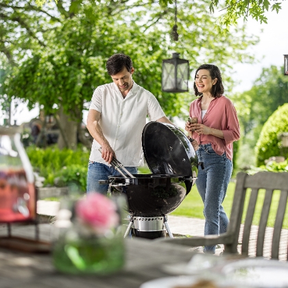 Sommerliche Grillszene im Garten: Ein Mann grillt am Kugelgrill, eine Frau steht neben ihm