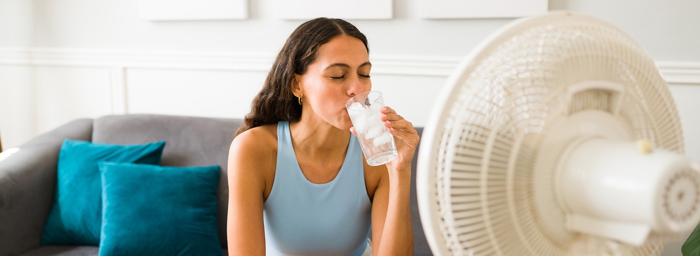 Frau sitzt auf dem Sofa vor einem Ventilator und erfrischt sich mit kaltem Wasser.