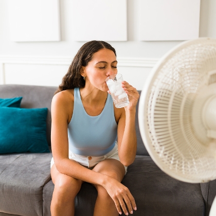 Frau sitzt auf dem Sofa vor einem Ventilator und erfrischt sich mit kaltem Wasser.