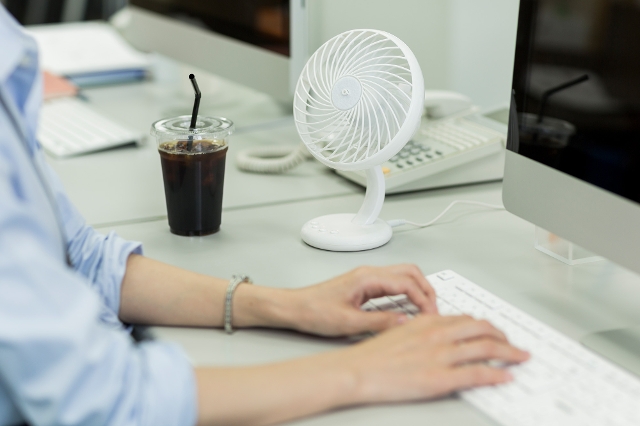 Eine Frau sitzt im Büro am Schreibtisch, auf dem ein Tischventilator für Abkühlung sorgt.